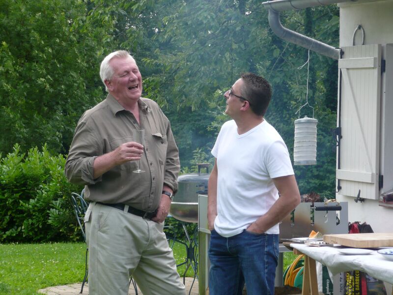 Two men laughing with glasses in hand beside the barbecue