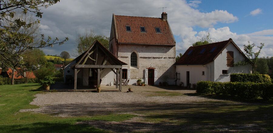 Le Verger from the drive: the timber-framed covered terrace, the white stone chapel and the west wing under a blue sky