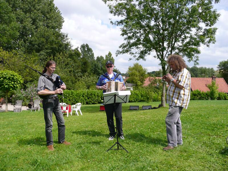 Three musicians playing on the lawn, accordion and pipes, with tables behind them