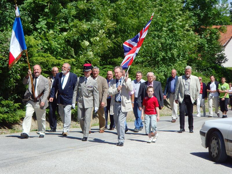 Men walking down a village lane behind a French flag and a Union flag, a boy in a red shirt among them