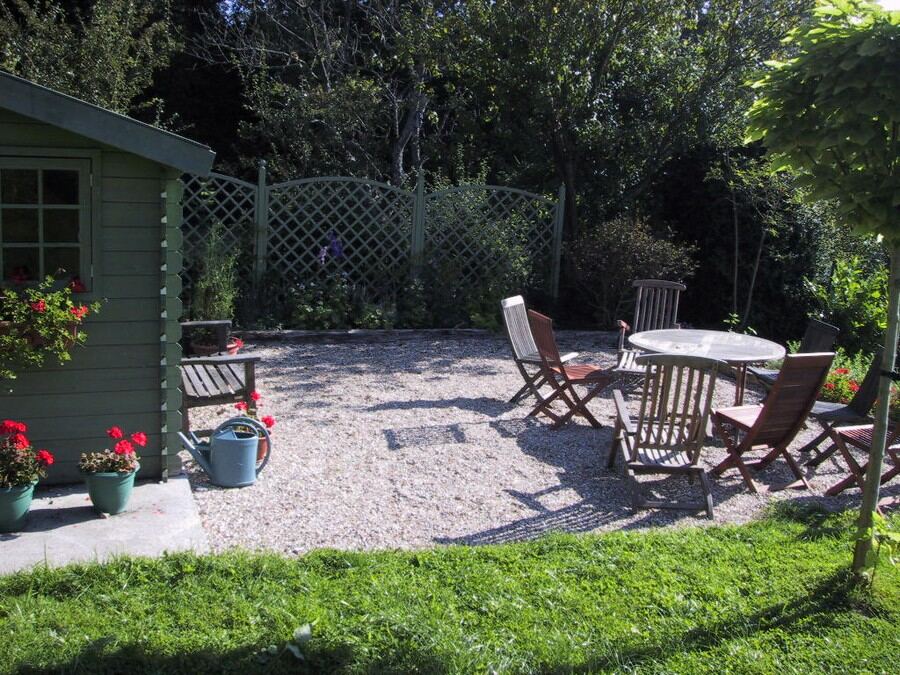 A gravel patio with a wooden table and chairs beside the green shed and trellis